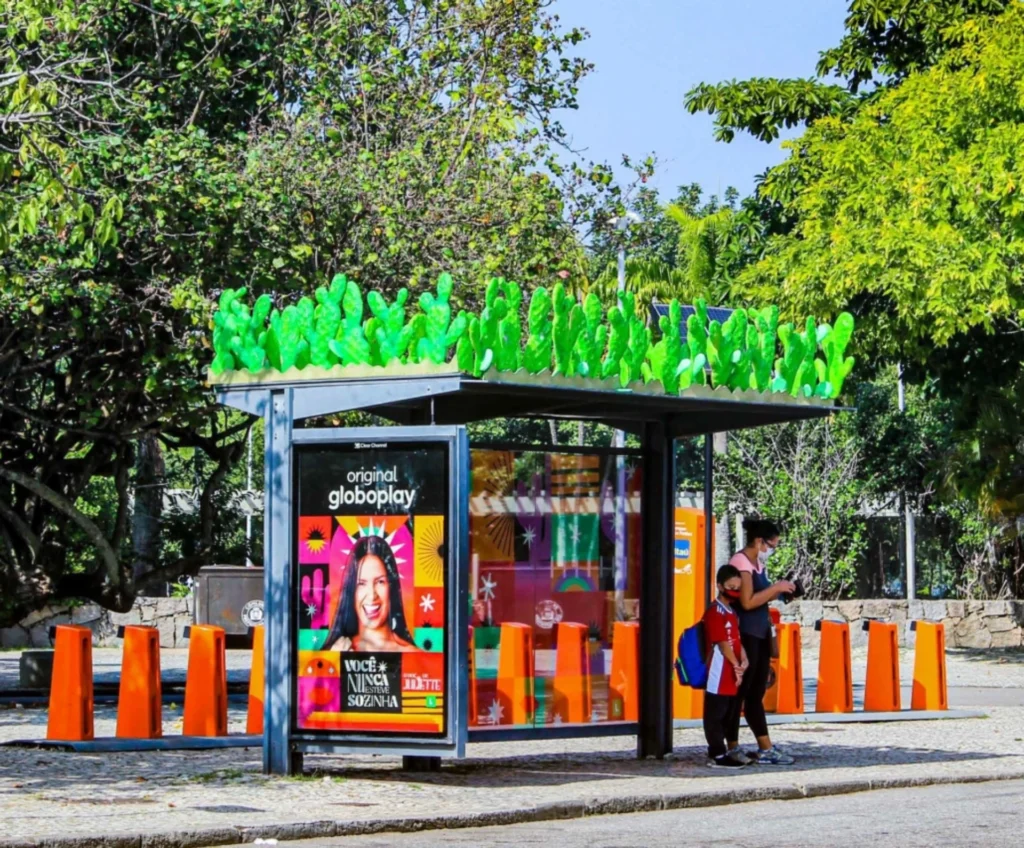 Parada de ônibus em uma área arborizada, decorada com plantas artificiais no teto, exibindo um anúncio da Globoplay. Pessoas aguardam sob a estrutura moderna, que traz um toque vibrante com cores e vegetação simulada.